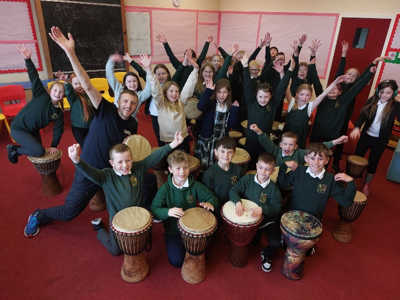 Children and music tutor raising hands beside drums in a music class.