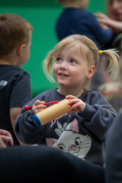 Young child holding a guiro percussion instrument, with other children in the background.  Happy Little Tunes