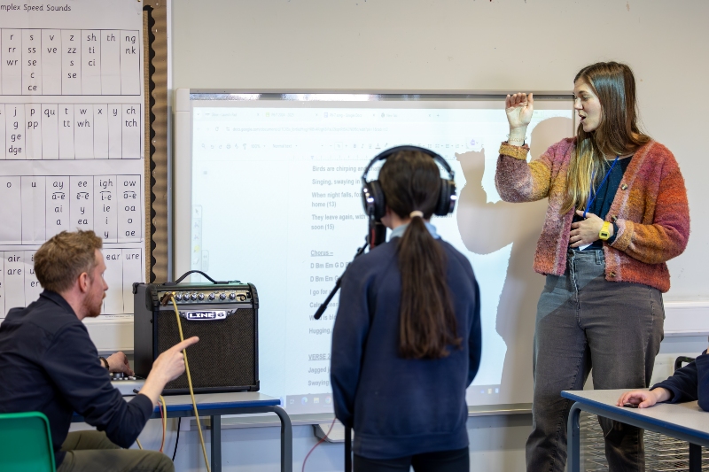 Classroom scene with three people: one pointing at an amplifier, one presenting near a whiteboard, and a pupil wearing headphones. A phonics chart with sounds like “sh,” “ch,” and “th” is visible.