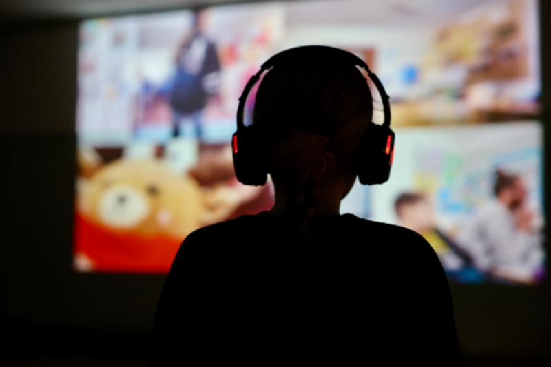 Silhouetted young person with headphones facing a screen of blurred video call images.