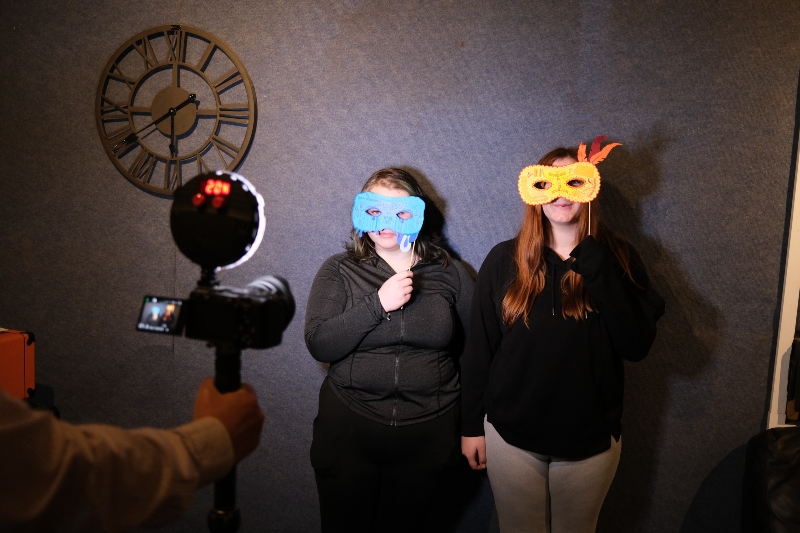 Two people with coloured masks covering their faces stand in front of a dark wall with a large clock, while someone films them with a tripod-mounted camera.
