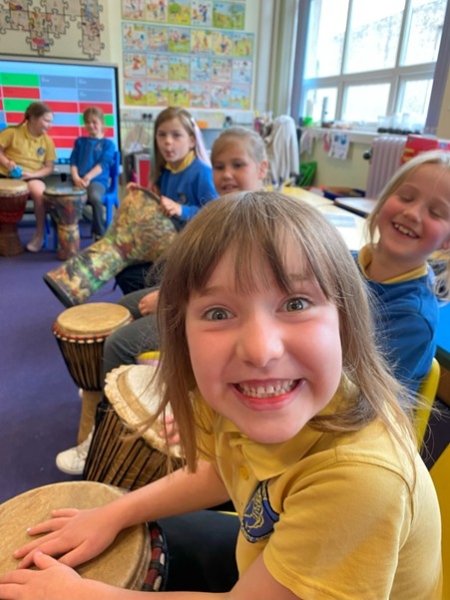 Children in school uniforms playing drums in a classroom. The one in the front has a huge grin.