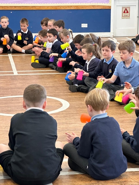 Children sitting in a circle on a wooden floor, holding colourful plastic cups used as an instrument during a group activity.