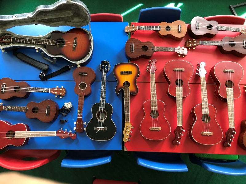 15 assorted ukuleles and small on blue and red tables, with colourful chairs around them.