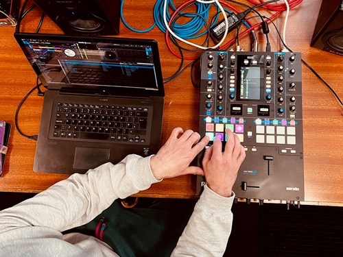 Young person using a Pioneer DJ controller and laptop with DJ software, surrounded by colorful cables on a wooden table.