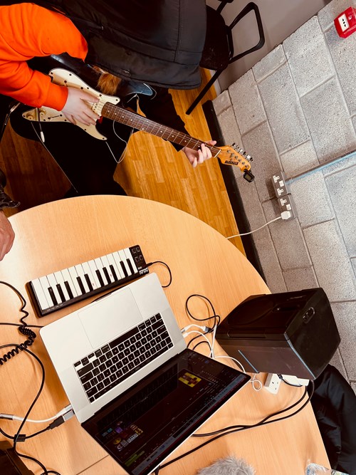 Young person playing an electric guitar with a small keyboard, laptop and speaker on the table in front of them.