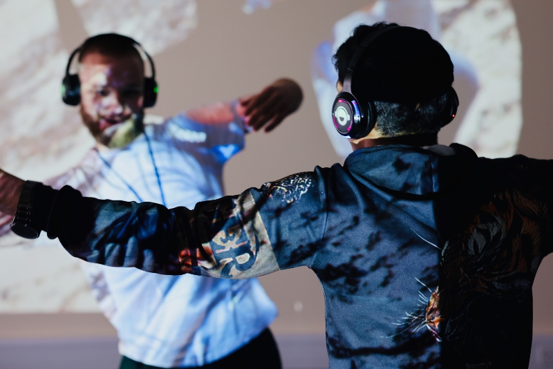 Two people wearing headphones, seen from behind with arms outstretched, in front of a colourful abstract projection.